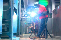 A pipefitter performs electric arc welding to attach a fitting.